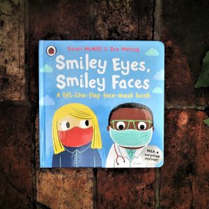 A board book: Smiley Eyes, Smiley Faces. Two people's faces are on the front, a Black doctor and a white woman with blonde hair. Both are wearing protective face masks.